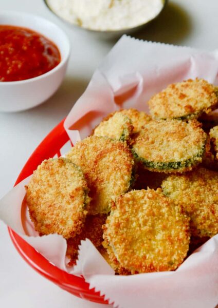 Baked Parmesan Zucchini Chips in red basket lined with white food paper with ketchup and mayonnaise in the background.