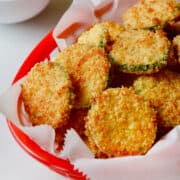 A paper-lined red food basket containing Baked Parmesan Zucchini Chips next to small bowls filled with marinara sauce and Parmesan cheese.