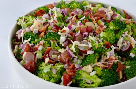 White bowl containing The Best Broccoli Salad with Bacon including crumbled bacon, broccoli florets, sliced almonds, red grapes, shredded mozzarella cheese and red onion.