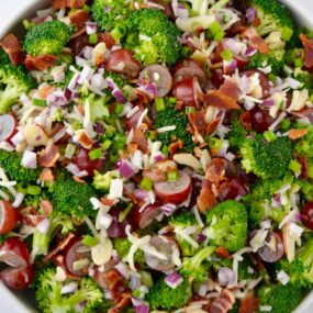 Large white bowl containing The Best Broccoli Salad with Bacon including crumbled bacon, broccoli florets, sliced almonds, red grapes, shredded mozzarella cheese and red onion.