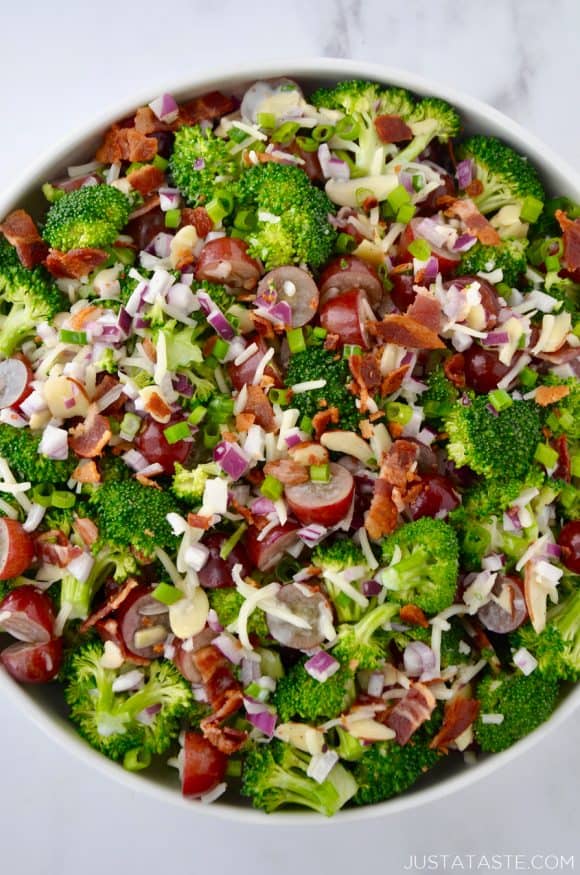 Large white bowl containing The Best Broccoli Salad with Bacon including crumbled bacon, broccoli florets, sliced almonds, red grapes, shredded mozzarella cheese and red onion.