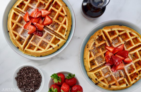 Two pale blue plates with Buttermilk Chocolate Chip Waffles topped with strawberries and powdered sugar surrounded by a small glass pitcher of maple syrup, and two small white bowls containing mini chocolate chips and fresh strawberries.