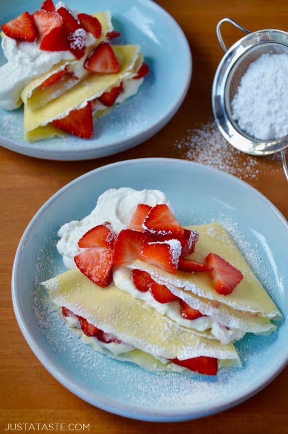 Two pale blue plates with Strawberry Cream Cheese Crêpes topped with fresh strawberries and whipped cream dusted with powdered sugar.