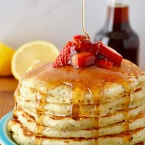 Lemon Poppyseed Pancakes topped with strawberries and maple syrup on blue plate with half a lemon and maple syrup in background.