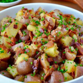 White bowl with Potato Salad with Warm Bacon Dressing next to small white bowl filled with fresh chives and wooden serving spoon and fork.