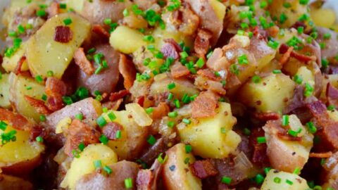 White bowl with Potato Salad with Warm Bacon Dressing next to small white bowl filled with fresh chives and wooden serving spoon and fork.