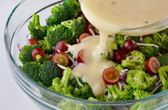 Broccoli salad dressing being poured into a glass bowl with broccoli florets, red grapes, diced red onion and shredded mozzarella cheese.