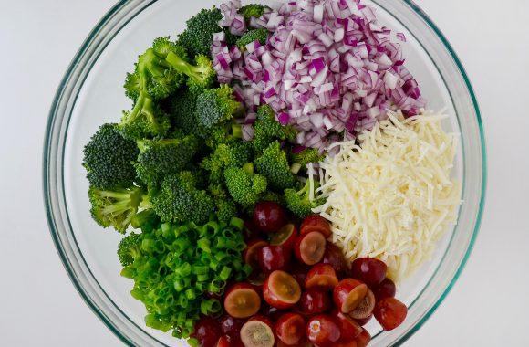 Glass bowl containing diced red onion, hredded mozzarella cheese, halved red grapes, chopped scallions and broccoli florets. 