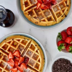 Buttermilk Chocolate Chip Waffles topped with strawberries and powdered sugar on pale blue plates next to a small white bowl filled with chocolate chips, a small glass pitcher with maple syrup and a small white bowl with strawberries.