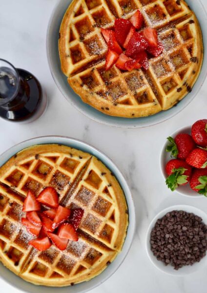 Buttermilk Chocolate Chip Waffles topped with strawberries and powdered sugar on pale blue plates next to a small white bowl filled with chocolate chips, a small glass pitcher with maple syrup and a small white bowl with strawberries.