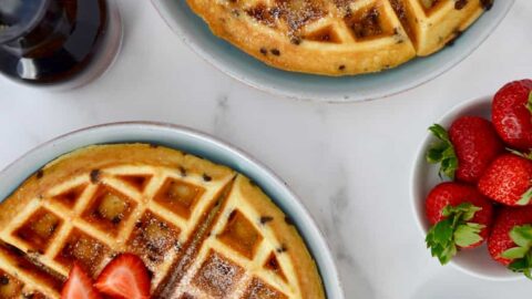 Buttermilk Chocolate Chip Waffles topped with strawberries and powdered sugar on pale blue plates next to a small white bowl filled with chocolate chips, a small glass pitcher with maple syrup and a small white bowl with strawberries.