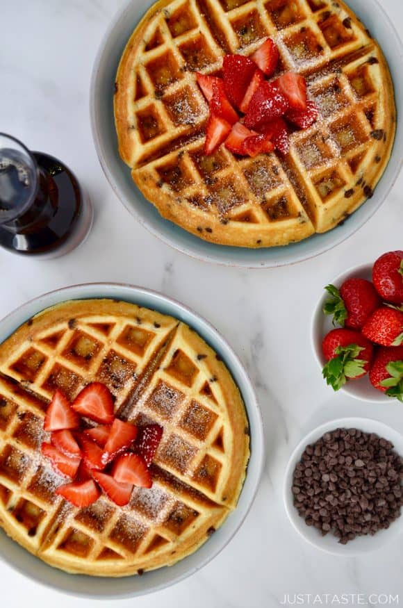 Buttermilk Chocolate Chip Waffles topped with strawberries and powdered sugar on pale blue plates next to a small white bowl filled with chocolate chips, a small glass pitcher with maple syrup and a small white bowl with strawberries. 