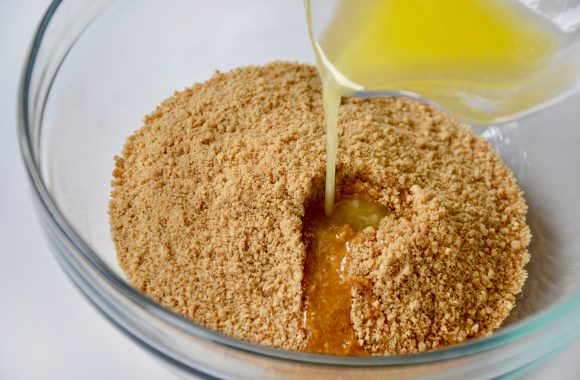 Crushed graham crackers in glass bowl with melted butter being poured on top. 