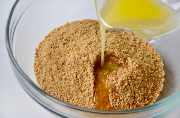 Crushed graham crackers in glass bowl with melted butter being poured on top.