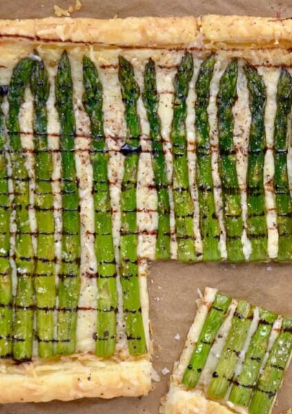 Cheesy Asparagus Tart drizzled with balsamic syrup with corner cut into two pieces on brown paper.