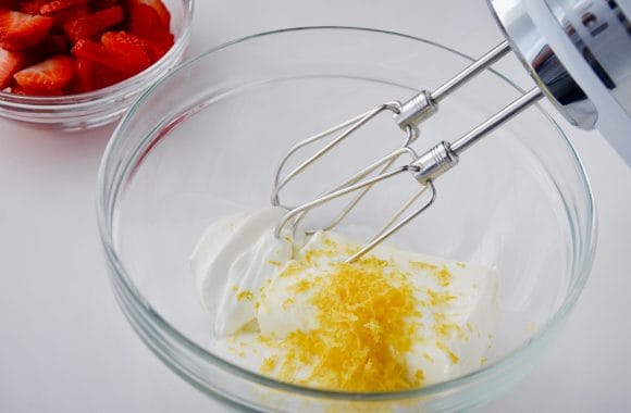 Glass bowl containing cream cheese, sour cream, sugar and lemon zest with handheld beater resting against it and small glass bowl with fresh strawberries in background. 