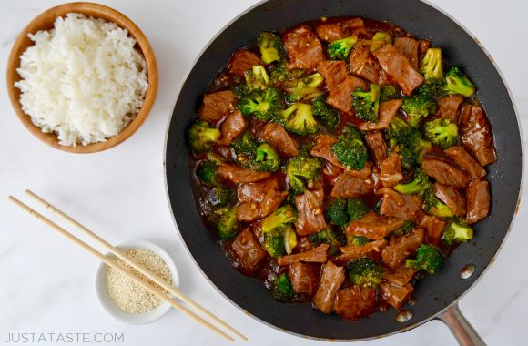 Easy Beef and Broccoli in a nonstick sauté pan, white rice in a small brown bowl and sesame seeds in a small white ramekin with chopsticks resting on top.