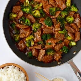 Easy Beef and Broccoli in a nonstick sauté pan, white rice in a small brown bowl and sesame seeds in a small white ramekin with chopsticks resting on top.