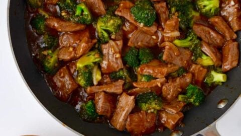 Easy Beef and Broccoli in a nonstick sauté pan, white rice in a small brown bowl and sesame seeds in a small white ramekin with chopsticks resting on top.