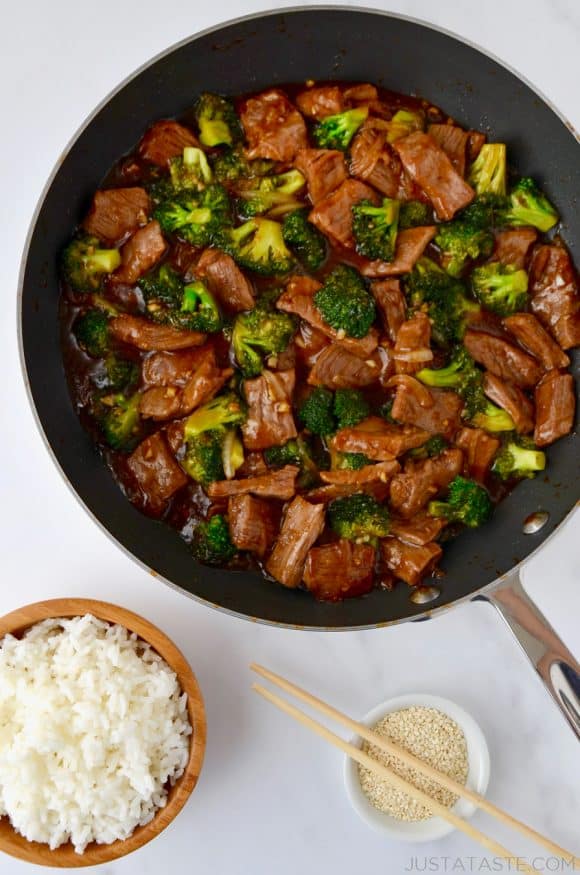 Easy Beef and Broccoli in a nonstick sauté pan, white rice in a small brown bowl and sesame seeds in a small white ramekin with chopsticks resting on top.