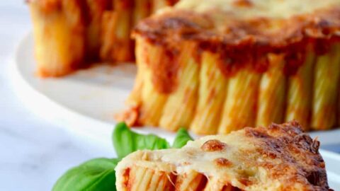 Slice of Easy Cheesy Rigatoni Pie on white plate with basil garnish; Easy Cheesy Rigatoni Pie in background with two glasses of wine and basil beside it