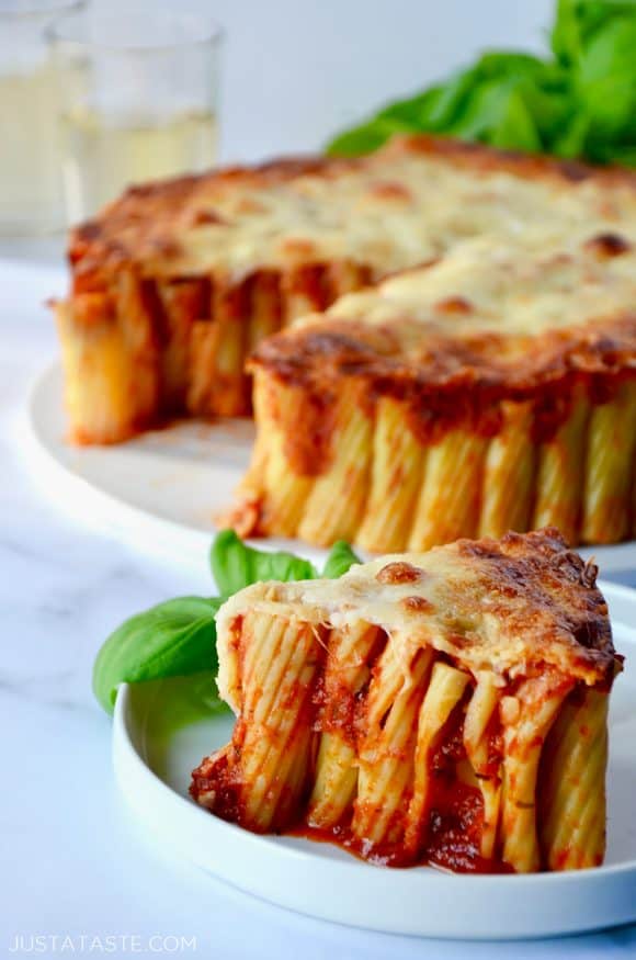 Slice of Easy Cheesy Rigatoni Pie on white plate with basil garnish; Easy Cheesy Rigatoni Pie in background with two glasses of wine and basil beside it