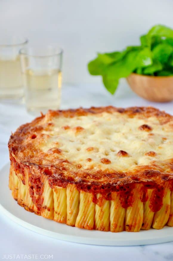 Easy Cheesy Rigatoni Pie on white serving plate with two glasses filled with wine and small wooden bowl with basil in background