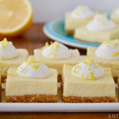 Square-cut Easy Lemon Cheesecake Bars on white plate with cut lemon and more bars on a blue plate in background.