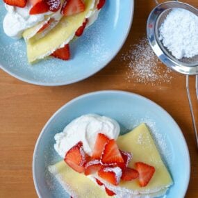 Two pale blue plates with Strawberry Cream Cheese Crêpes topped with fresh strawberries, whipped cream and powdered sugar.