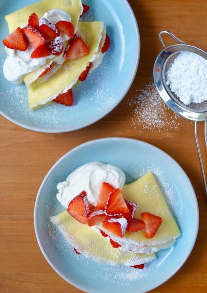 Two pale blue plates with Strawberry Cream Cheese Crêpes topped with fresh strawberries, whipped cream and powdered sugar.