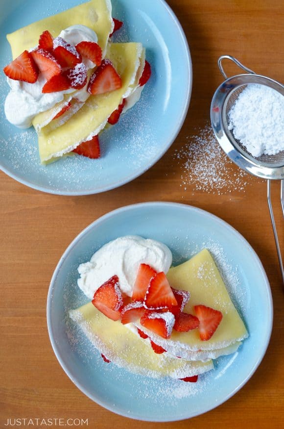 Two pale blue plates with Strawberry Cream Cheese Crêpes topped with fresh strawberries, whipped cream and powdered sugar.