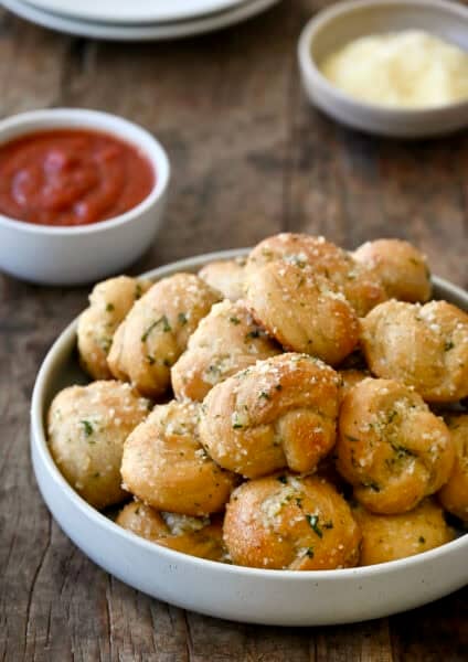 Homemade garlic knots made with pizza dough, brushed with garlic butter and topped with grated Parmesan and chopped fresh parsley in a serving bowl.