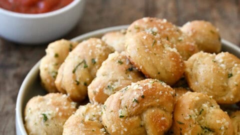 Homemade garlic knots made with pizza dough, brushed with garlic butter and topped with grated Parmesan and chopped fresh parsley in a serving bowl.