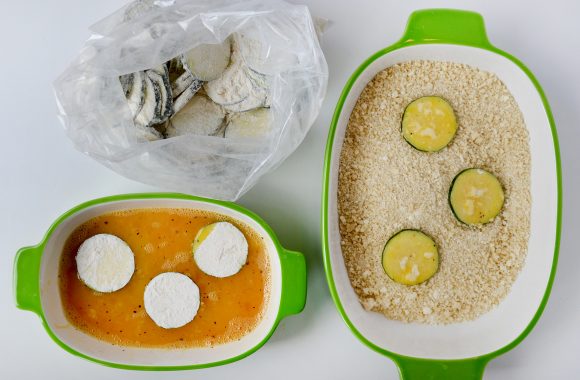 Bag containing flour-coated zucchini, small white and green bowl containing egg mixture and three flour-coated zucchini chips, and one large white and green bowl containing breadcrumbs and three zucchini chips.