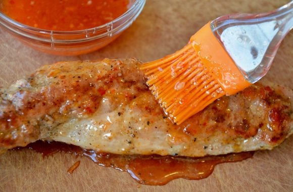 Roast pork tenderloin on cutting board being brushed with Thai sweet chili sauce with more chili sauce in small clear bowl in the background.