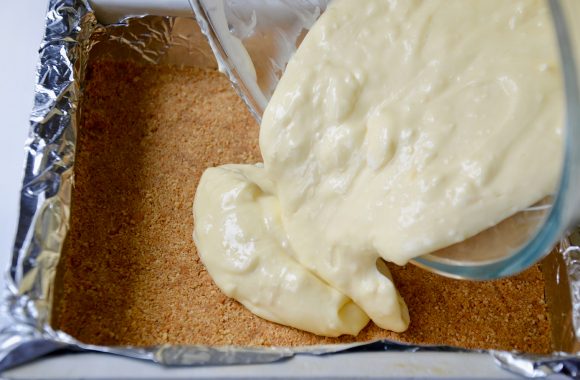 Lemon cheesecake mixture being poured over graham cracker crust in a aluminum foil lined square baking pan. 