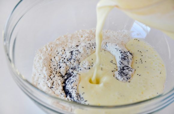 Glass bowl with flour, sugar, baking soda, baking powder, poppy seeds and salt with wet ingredients being poured into bowl.