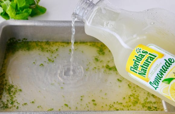 A bottle of Florida's Natural Lemonade being poured into a metal baking pan filled with chopped up mint leaves
