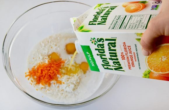 A glass bowl containing eggs, orange zest and coconut milk with orange juice being poured in