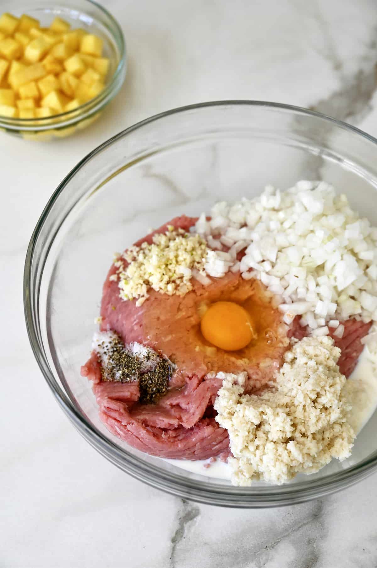 A glass bowl containing ground pork, breadcrumbs, diced onion, an egg, salt, pepper and minced garlic.