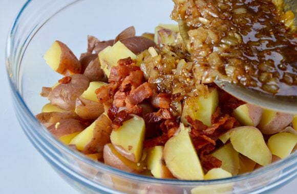 Glass bowl with quartered red potatoes and cooked bacon with warm bacon dressing being poured on top. 