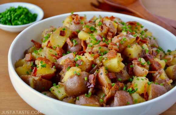 White bowl with Potato Salad with Warm Bacon Dressing garnished with fresh chives with small white bowl filled with fresh chives and wooden serving utensils in background.