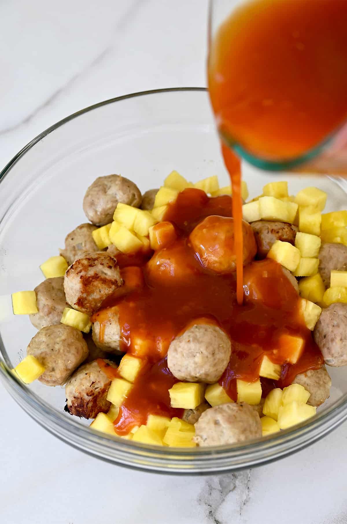 Sweet and sour sauce being poured over meatballs and pineapple chunks in a glass bowl.
