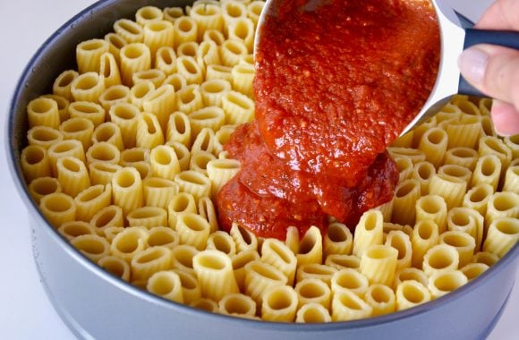 Rigatoni standing up in 9-inch springform pan with marinara sauce being poured on top from measuring cup