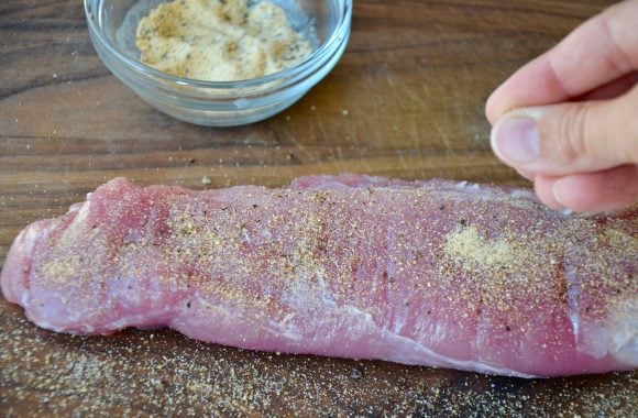Pork tenderloin being seasoned with small clear bowl containing seasonings in background.