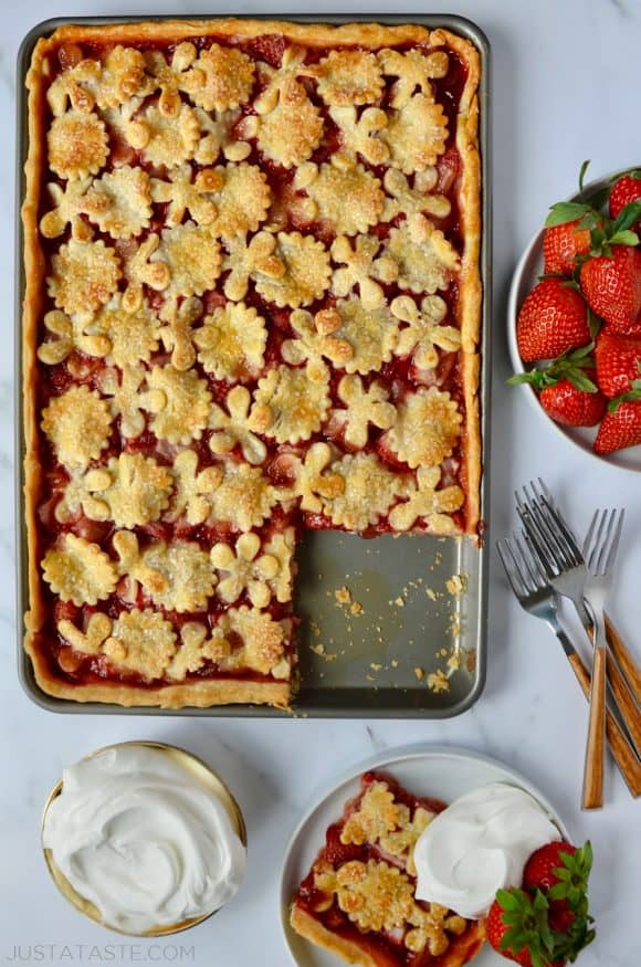 Simple Strawberry Slab Pie with a single serving on a white plate, whipped cream, forks and a bowl of fresh strawberries surrounding it.