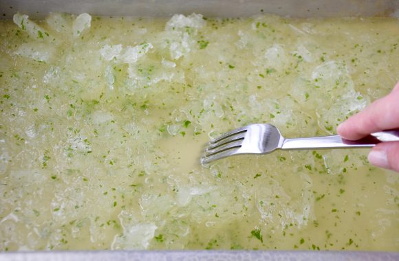 A fork stirring a pan of lemon granita made with chopped up mint leaves