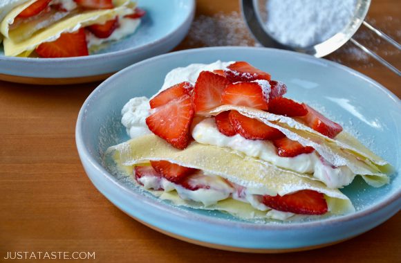 Strawberry Cream Cheese Crepes topped with fresh strawberries and whipped cream dusted with powdered sugar on pale blue plate with mesh sifter containing powdered sugar in background.
