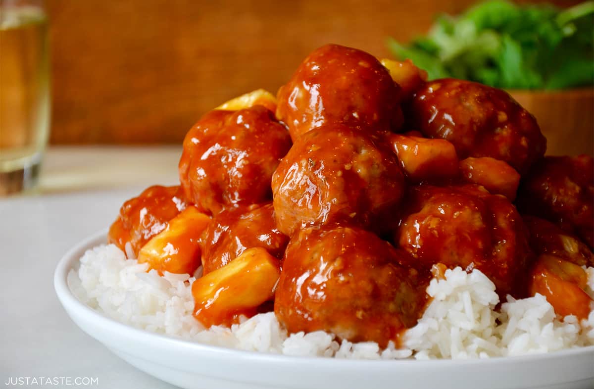 Baked sweet and sour meatballs with pineapple chunks over white rice in a white dinner bowl.