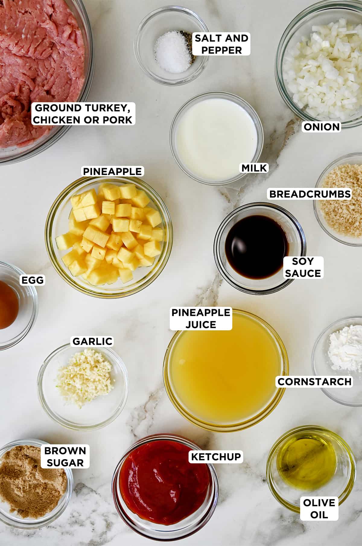 Various sizes of glass bowls containing the ingredients needed to make meatballs with a sweet and sour pineapple sauce, including ground pork, milk, diced onion, Panko breadcrumbs, pineapple juice, soy sauce, pineapple chunks, minced garlic, egg, cornstarch, olive oil, brown sugar and ketchup.